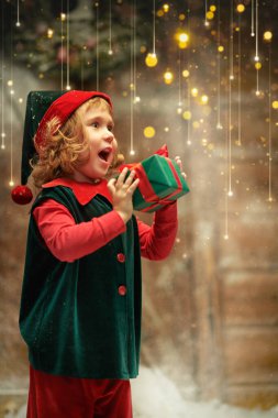 A portrait of a little elf girl with cute curly hair, who stands by a wooden hut holding a gift in her hands and joyfully laughing. Magic Christmas time, presents. Christmas tale. 