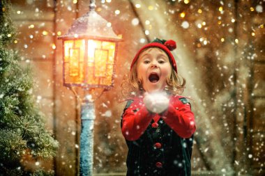 A cute little elf girl catches snowflakes and rejoices, standing at a wooden hut and a snow-covered lantern under a snowfall. Magic light flicker around. Winter Christmas tale. 