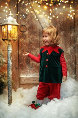 Magic Christmas time. A cute little  elf girl with curly blonde hair stands in the snowdrifts near a wooden hut  looking at the lantern with joy and admiration. Magic lights shine in the background.