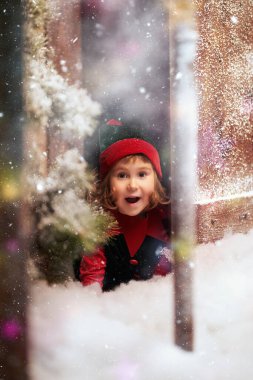 Time of Christmas miracles. Portrait of a cute little elf girl looking out excitedly from behind the door of a snow-covered wooden house in a fabulous winter forest.