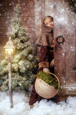 Full length shot of a joyful girl in medieval clothes bringing a basket of fir branches and fir cones from the forest into a wooden house. Historical reconstruction. Merry Christmas! 