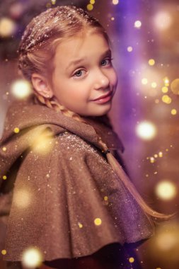 Time of Christmas miracles. A pretty little blonde girl with pigtails in winter medieval clothes standing at a wooden house surrounded by magical shining lights. Merry Christmas and Happy New Year! clipart