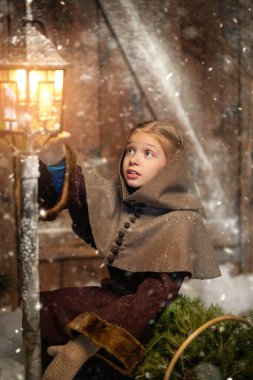 Old Christmas Tale. Cute little girl in medieval clothes sits at a snow-covered wooden house with a basket of spruce branches admiring the beauty of a burning lantern. 