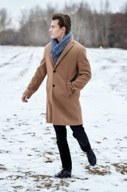 Full length shot of an elegantly-dressed young man in a stylish coat and scarf walking outdoor on a cold winter day. Men's beauty and fashion.
