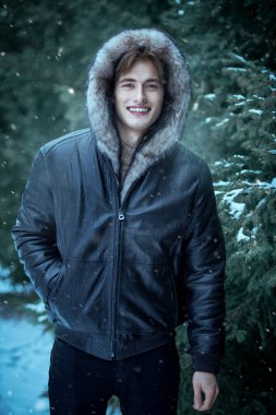 Attractive young man in stylish black leather jacket with a fur hood on his head poses in the park cheerfully smiling in the background of fir trees on a cold, snowy day. Men's winter fashion.