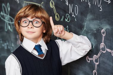 Education. Portrait of a cute smart boy in neat school uniform wearing glasses standing at the blackboard with chemical formulas and holding his index finger up. Clever kids.  