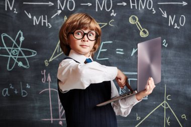 Portrait of an excited clever little boy in school uniform and glasses pressing keys on his laptop in the background of a blackboard with scientific formulas. Smart children. Education. 