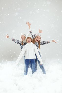 Kid's winter fashion. Three cute, cheerful children in stylish knitted sweaters and hats pose in a snowy setting joyfully screaming and streching their arms aside. Studio portrait. Winter activities.