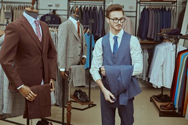 Zarif klasik takım elbiseli ve gözlüklü yakışıklı bir esmer, şık erkek giyim mağazasından kıyafet seçiyor. Erkeklerin iş modası. 