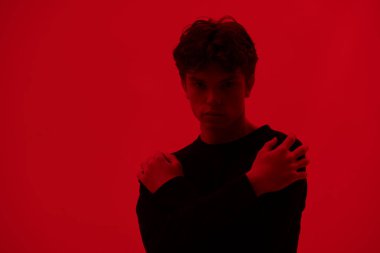 Kollarını saran dalgalı siyah saçlı depresif yakışıklı bir adamın portresi. Stüdyo çekimi koyu kırmızı arka planda. Gençler ve akıl sağlığı.