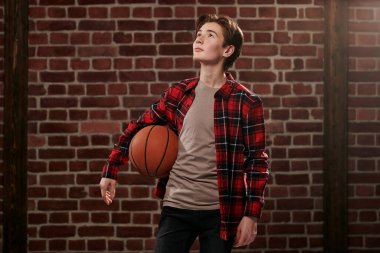 Modern bir genç, gündelik giysiler içinde elinde bir basketbol topuyla tuğla duvara dayanıyor. Spor, hobiler. Etkin yaşam biçimi.