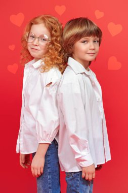 Beyaz gömlekli ve kotlu neşeli kız ve oğlan sırt sırta durup kameraya bakıyor. Çocuklar, çocukluk, moda ve eğitim. Kırmızı arka planda kalpleri olan bir stüdyo resmi.