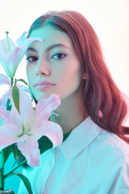 Güzellik ve çiçekler. Parlayan beyaz bir arka planda narin zambak çiçekleri olan güzel kahverengi saçlı bir kızın portresi. Makyaj ve makyaj malzemeleri. Parfüm. 