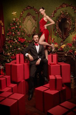 Noel ve yeni yıl modası. Lüks gece elbiseleri içinde yakışıklı bir adam ve güzel bir kız Noel için süslenmiş pahalı iç dekorasyonlarda kırmızı hediye kutularının arasında poz veriyorlar. Tatil indirimi ve alışveriş konsepti. 