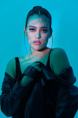 Parlak akşam makyajı ve parlak neon yazıtları olan esmer bir kızın atölye çekimi. Yüzüne yansıyan parlak, deri ceketli, mavi bir arka plan. Güzellik, makyaj ve kozmetik konsepti. 