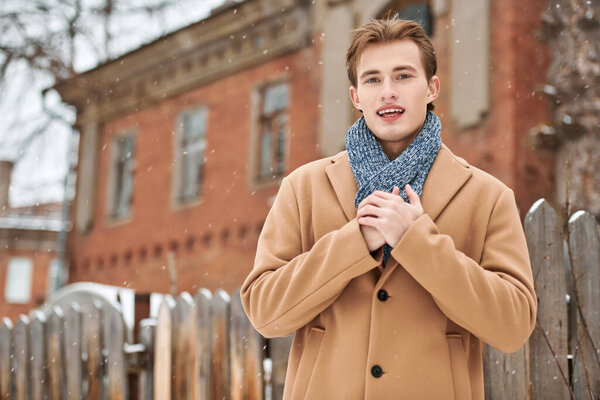 Joyful handsome young man in a trendy coat and scarf standing on the city street by a brick house on a cold winter day. Men's fashion and lifestyle.