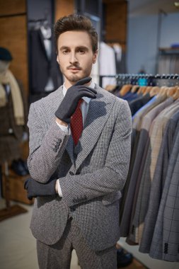 Yakışıklı, üç parçalı eldivenli şık takım elbiseli esmer adam erkek giyim mağazasında yeni kıyafetler seçiyor. Erkeklerin iş modası.