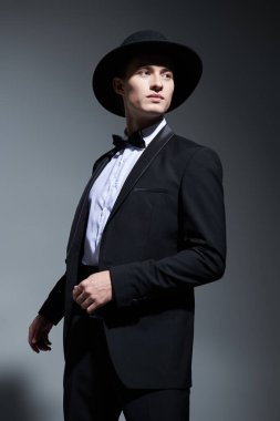 Gri bir stüdyo arka planında poz veren şık siyah takım elbiseli, papyonlu ve şapkalı genç yakışıklı bir adam. Erkek modası ve güzelliği. 