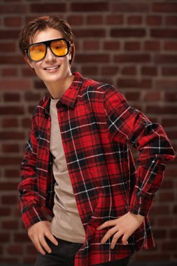 Modern bir gencin portresi şık bir gençlik giysisi ve güneş gözlüğü takmış tuğla duvarın arkasında neşeyle gülümsüyor. Gençlik tarzı. Moda..