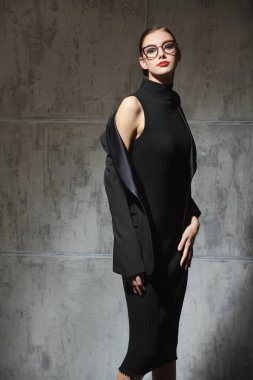 İş gündelik tarzı. Akşam makyajı ve şık gözlükleri olan zarif bir kadının portresi. Siyah örgü bir elbise ve grunge bir arka planda siyah bir ceket giyiyor. Moda fotoğrafı.. 