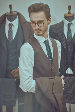 Portrait of an attractive young brunet man dressed in a smart brown three-piece suit standing in a men's clothing store. Men's business fashion. clipart