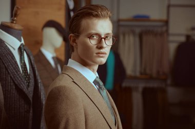 Zarif kahverengi takım elbiseli ve gözlüklü yakışıklı sarışın bir adamın portresi. Premium erkek giyim mağazasında duruyor. Erkeklerin iş modası. 