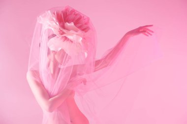 Kafasında büyük bir şakayık çiçeğiyle şeffaf organzanın altında açık pembe bir arka planda poz veren güzel, sofistike bir kızın stüdyo fotoğrafı. Deri ve vücut bakımı. Kozmetik ve parfüm. 