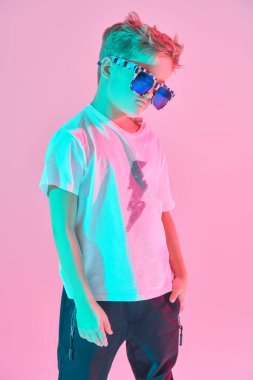 Beyaz tişörtlü ve şık güneş gözlüklü şık bir gencin portresi. Pembe arka planda pembe ışıklı bir stüdyo resmi. Çocuk modası..