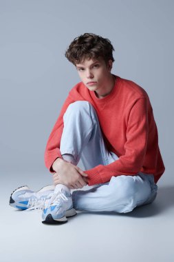 Kırmızı kazaklı yakışıklı adam modeli, açık kot pantolon ve stüdyoda yerde oturan spor ayakkabıları. Erkeklerin gençlik modası. 