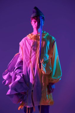 Gençlik ve parlak renkler. Havalı adam parlak sarı bir ceket ve parlak mor saçlı yeşil bir şapka takıyor. Pembe ışıkta mor bir arkaplanda stüdyo çekimi. Yaratıcı tasarımcı giysileri. 