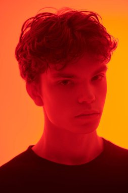 Kırmızı ışıklandırmalı, turuncu arka planda duran kapkara saçlı yakışıklı bir adamın stüdyo fotoğrafı. İnsanlar, duygular. 