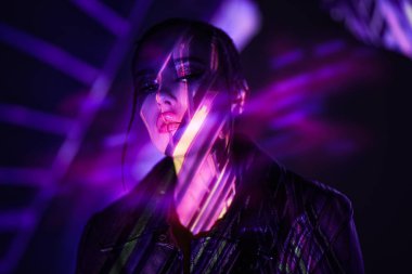 Gelecekçi stil, güzellik ve moda. Siyah deri ceket giyen çekici esmer kız yüzünde neon ışıklı ışıklarla karanlık bir alanda poz veriyor. Sanal gerçeklik kavramı. 