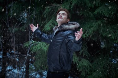 Parkta kürklü siyah deri ceketli genç sarışın adam duruyor ve karlı bir kış gününde doğanın güzelliğine hayran. Erkek modası. Kış mevsimi. 