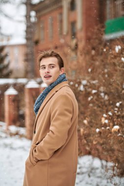 Erkek güzelliği, moda ve yaşam tarzı. Şık bir palto ve eşarp giymiş neşeli yakışıklı bir genç adam karlı bir günde sokakta yürüyor ve birine bakıyor.. 