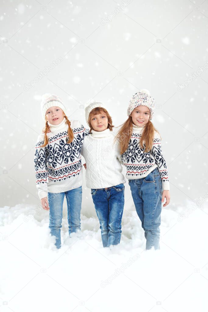 Children's winter fashion. Three cute, cheerful children in knitted sweaters, hats and jeans pose in a snowy setting. Full-length studio portrait. Winter activities.