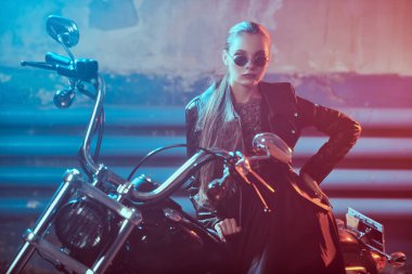 Siyah deri ceketli, siyah güneş gözlüklü çekici bir kız garajda motosikletine yaslanıyor..