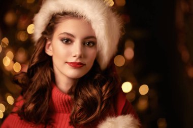 Kırmızı kazaklı ve Noel Baba şapkalı güzel bir kız Noel ağacının ve yanıp sönen ışıkların arkasına gülümsüyor. Noel ve Yeni Yıl Kutlaması.