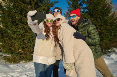 Bir grup modern genç kız ve erkek güneşli bir günde kış parkında dinlenip selfie çekiyorlar. Kış modası. İyi ruh hali, duygular. Kış aktiviteleri. 