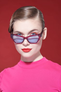 Parlak makyajlı ve gözlüklü zarif sarışın kız kırmızı arka planda poz veriyor. Makyaj ve makyaj malzemeleri. Gözlük stili. 