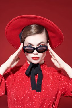 Moda ve güzellik. Parlak makyajlı çekici bir model kız kırmızı elbise, kırmızı şapka ve güneş gözlüğü takıyor. Kırmızı arka plan. Makyaj ve kozmetik. 