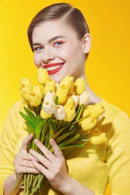 Parlak bahar renkleri. Parlak makyajlı sevimli bir kız elinde bir buket sarı laleyle sevinçle ayakta duruyor. Makyaj ve makyaj malzemeleri. Parlak sarı arkaplan.