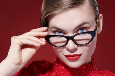 Gözlük stili. Parlak makyajlı ve zarif gözlüklü güzel bir kadın kırmızı arka planda poz veriyor. Makyaj ve kozmetik. 
