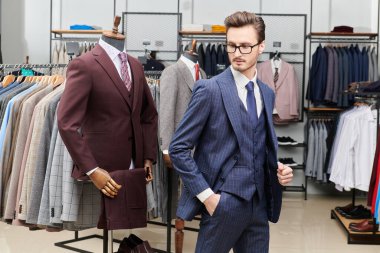 Gözlüklü, üç parçalı şık takım elbiseli yakışıklı bir iş adamı pahalı bir erkek giyim mağazasında lüks klasik takım elbiseler giyer. Erkek modası, iş tarzı.. 