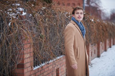 Tuğla evin kapısında duran ve gülümseyen şık ceketli ve eşarplı yakışıklı bir gencin portresi. Erkek güzelliği ve modası. Boşluğu kopyala.