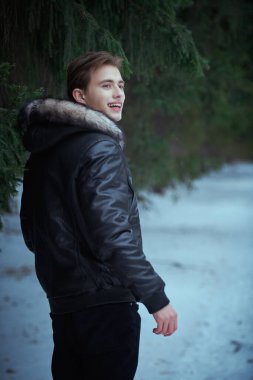 Erkekler kış modası. Yakışıklı, siyah deri ceketli genç bir adam parkta yürüyor köknar ağaçlarının ve yerde yatan karın arkasında neşeyle gülümsüyor.. 