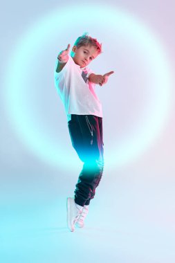 Modern dans stili. Parmak uçlarında dans eden havalı break dansçı çocuk stüdyoda parmaklarını beyaz arka planda karışık renkli ışıklandırmada kameraya doğrultuyor. Hip-hop dansçısı. Spor ve aktiviteler.