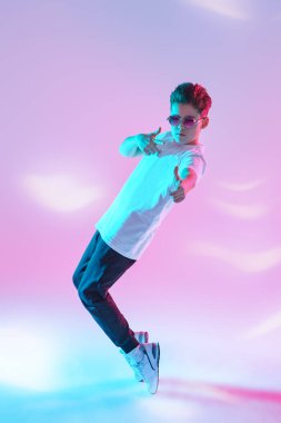 Çağdaş danslar. Profesyonel hip hop ve break dansçı çocuk dans ediyor ve parmaklarını stüdyoda pembe arka planda karışık renkli ışıkta kameraya doğrultuyor. Tam boy portre. 