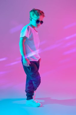 Çocuk modası. Moda gözlüklü, beyaz tişörtlü ve eşofmanlı genç bir çocuğun stüdyo portresi. Karışık ışık ile pembe arkaplan.