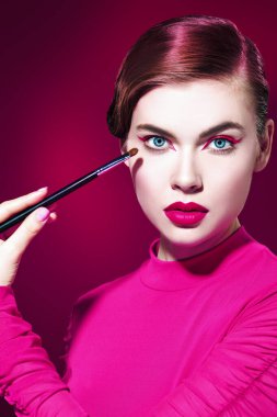 Parlak pembe rujlu ve gözlerinde pembe oklar olan güzel bir manken kız kırmızı arka planda bir fırçayla makyaj yapıyor. Güzellik, makyaj ve kozmetik. Makyaj araçları.