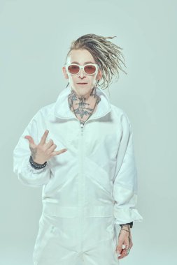 Gençlik alternatif kültürü. Beyaz takım elbiseli, beyaz güneş gözlüklü modern stil punk rock müzisyeni neşeyle gülümsüyor ve güzel bir jest yapıyor. Beyaz stüdyo geçmişi. 